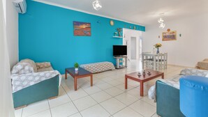 A living room with a blue wall, a sofa, a TV, a dining table, and a small plant. - Eftichia Nea Chora Apartment with Mountain View, Shared Garden & Wi-Fi (Chania)