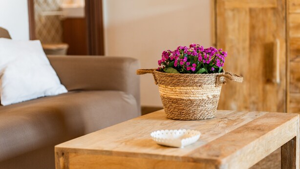 Een houten tafel met een geweven mand met paarse bloemen, een witte decoratieve schaal en een bruine leren bank op de achtergrond.