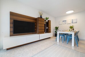 A modern living room with a flat-screen TV, wooden entertainment unit, dining table with chairs, and a patterned rug.