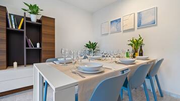 Un comedor con una mesa dispuesta para seis personas, rodeada de sillas, con una estantería y plantas en macetas al fondo.