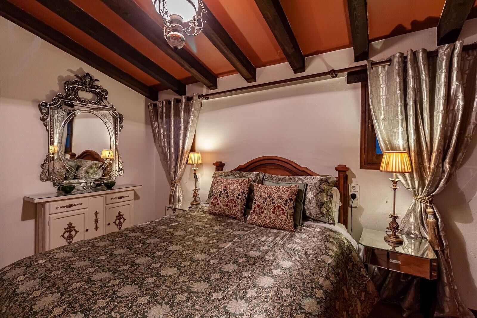 A bedroom with a patterned bedspread, a wooden headboard, a nightstand with a lamp, a dresser with a mirror, and curtains.
