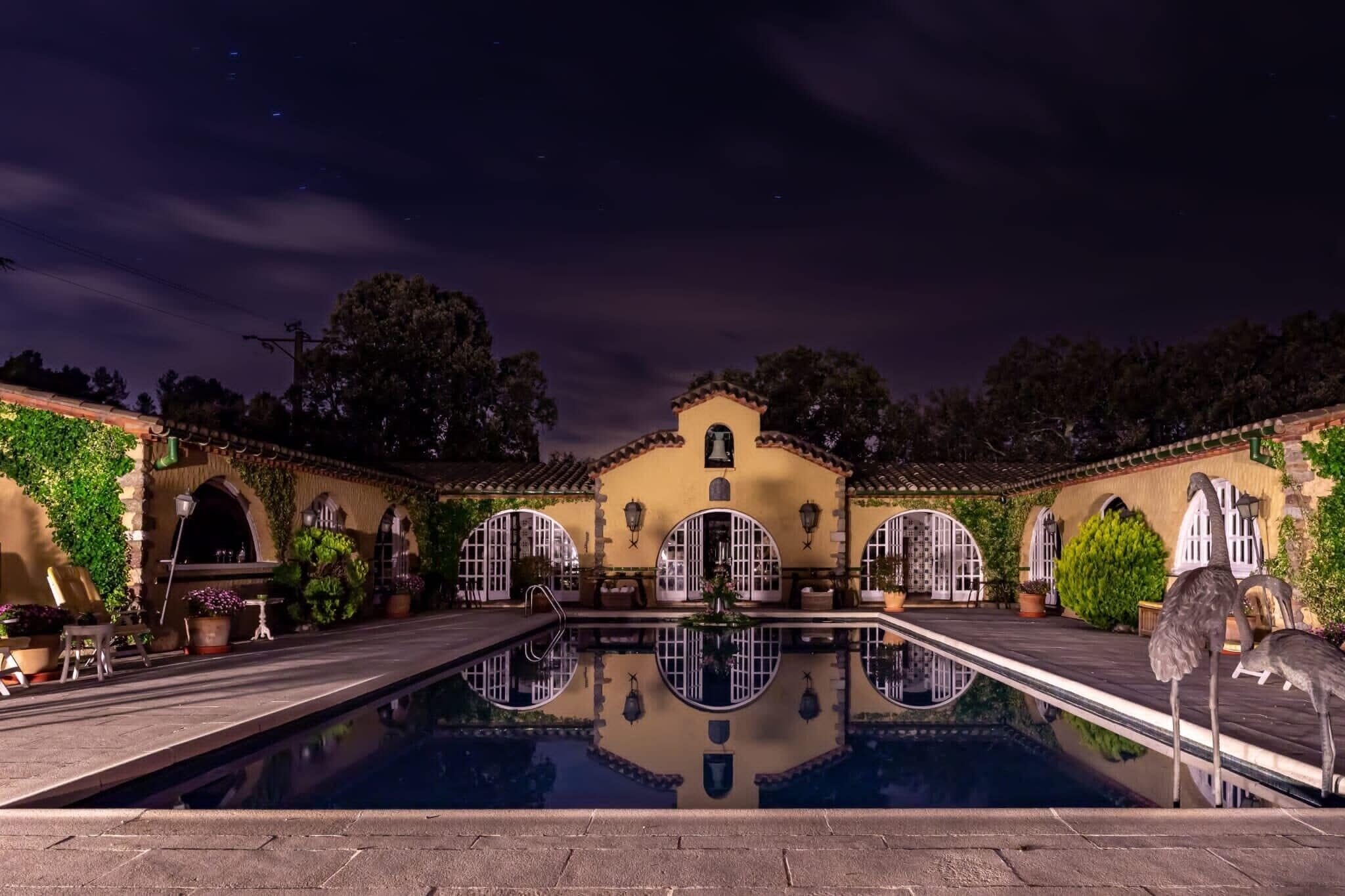 A nighttime pool area with a reflection, a building with arches, and a bird statue.