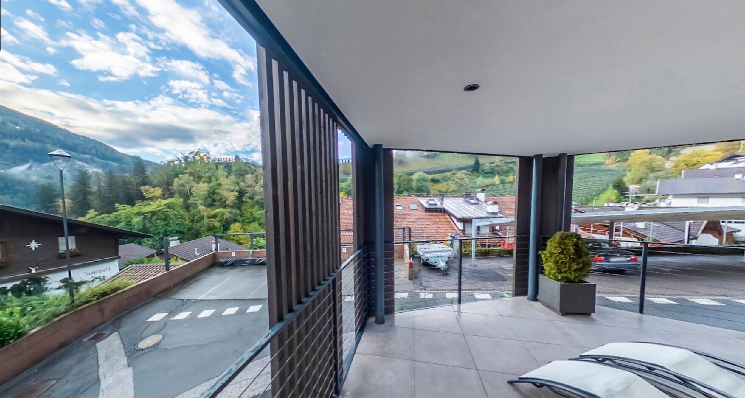 Un balcon donnant sur un paysage montagneux, un espace de stationnement et un bâtiment doté d’un toit en pente.