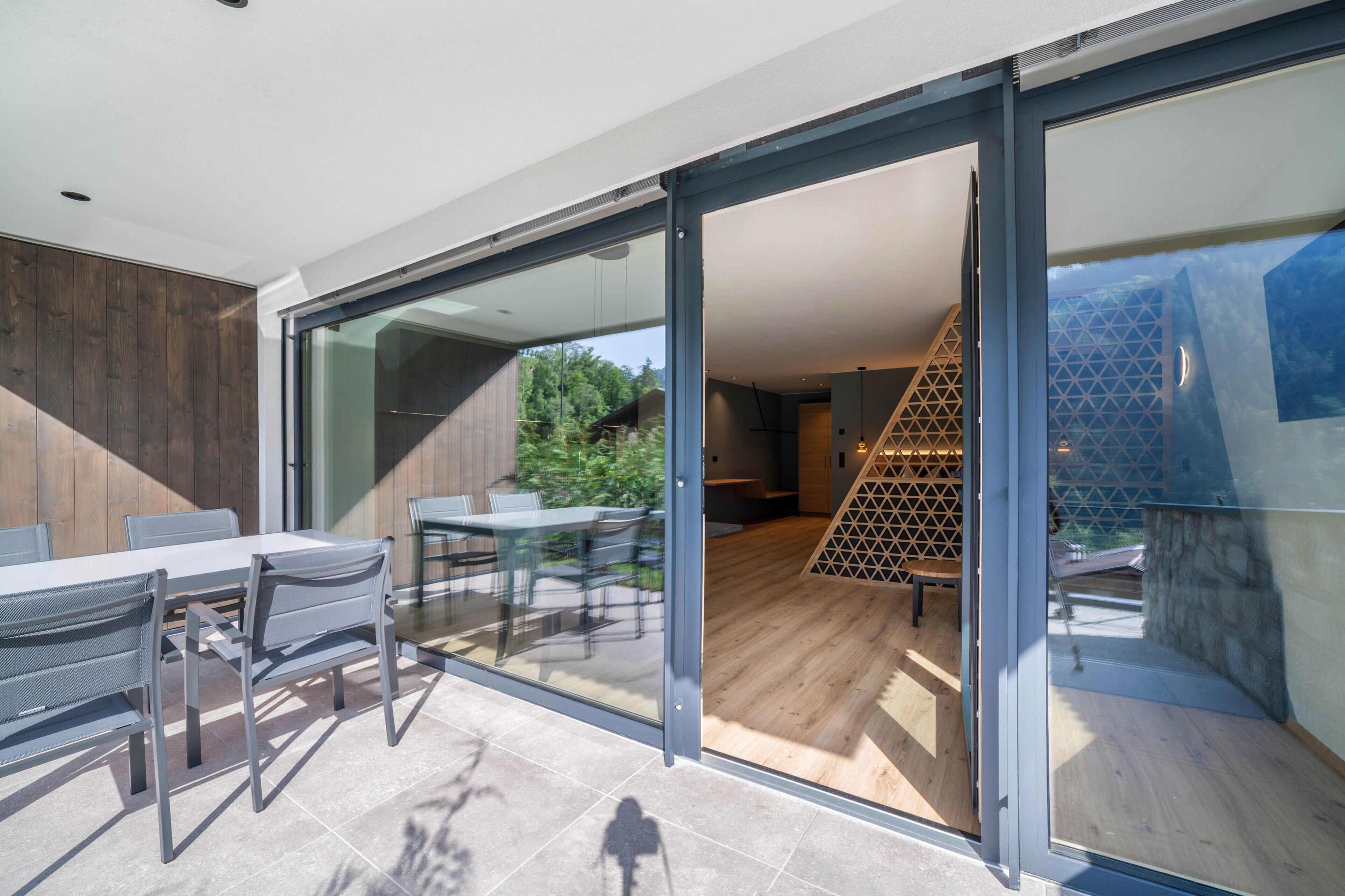 A modern outdoor dining area with a glass partition, a wooden deck, and a staircase leading to an upper level.
