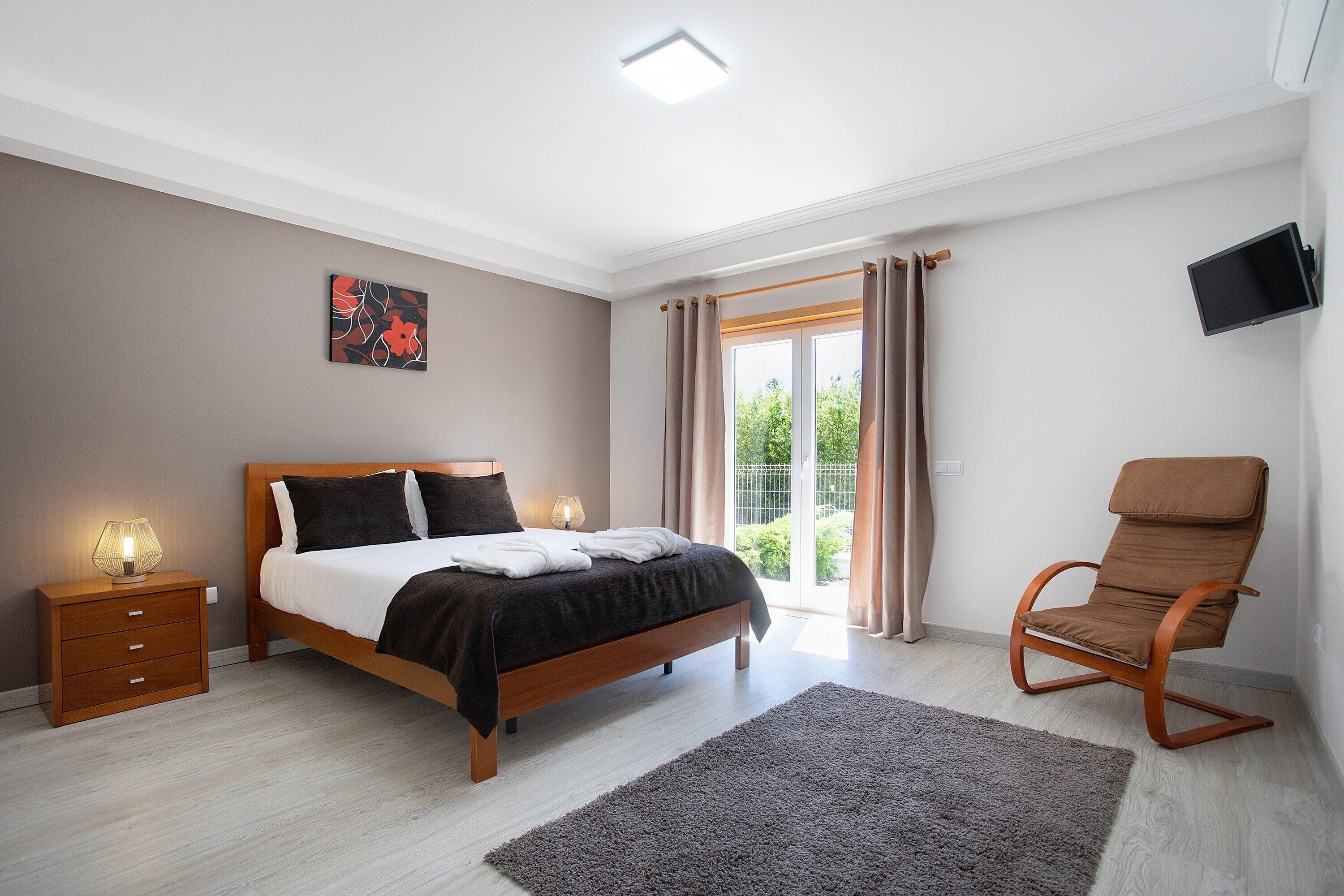 Un dormitorio con cama, mesita de noche, silla y televisor en la pared.