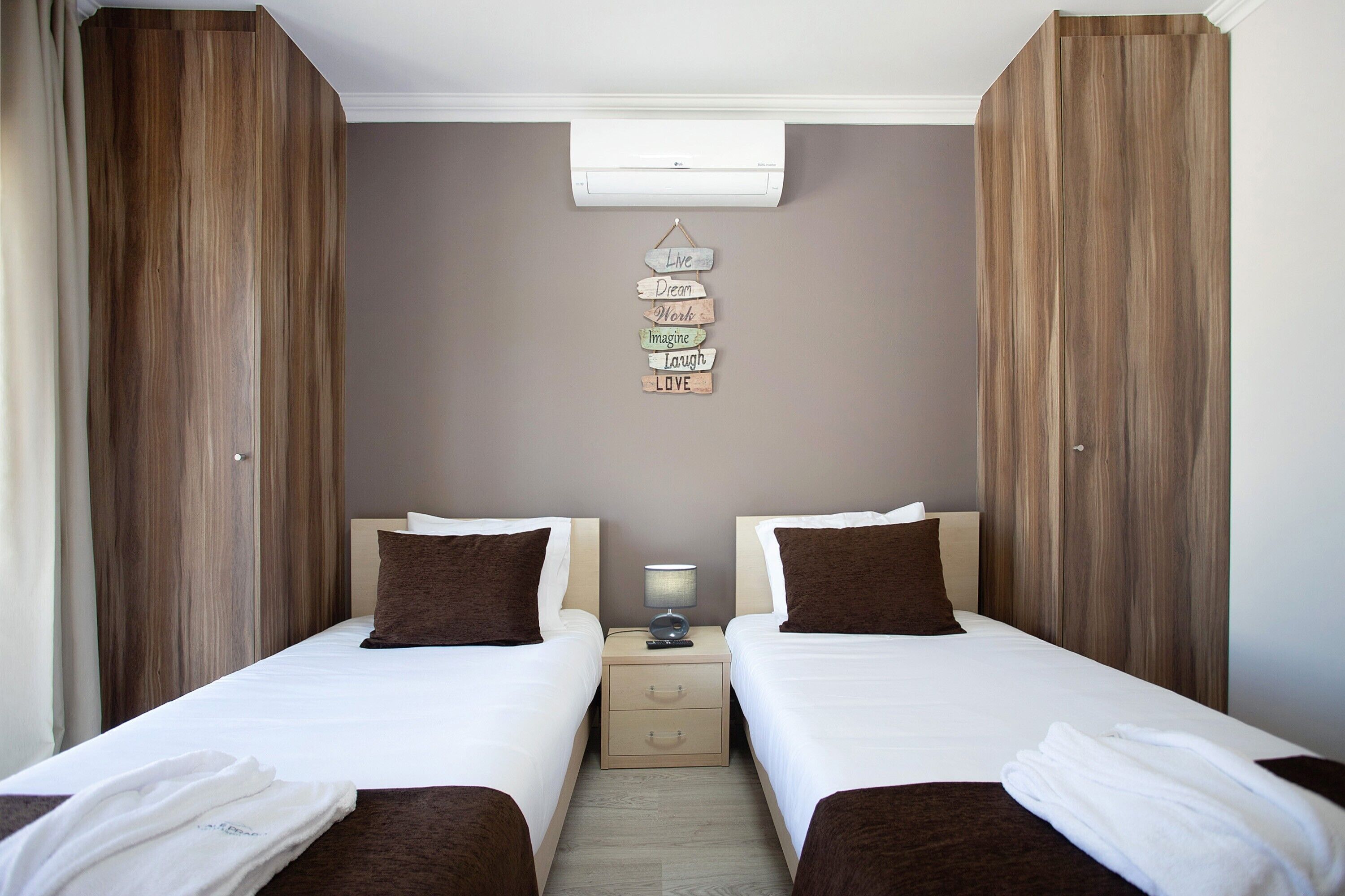 Une chambre d’hôtel avec deux lits, une table de chevet avec une lampe, un appareil de climatisation et une citation inspirante affichée sur le mur.