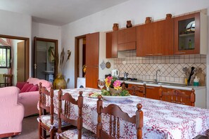 Zona de menjador amb taula preparada per a quatre, caires de fusta, cuina amb armaris de fusta i finestra amb cortines.