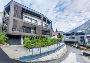 Un immeuble moderne à plusieurs étages, doté de balcons, d’un mur de soutènement en pierre et d’un espace paysager agrémenté de plantes.