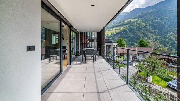 A balcony with a view of a mountainous landscape, a dining area with a table and chairs, and a glass door leading to an interior space. - Apartment "Lodge Terrace Exclusive Two" with Mountain View & Wi-Fi (Sankt Martin in Passeier)
