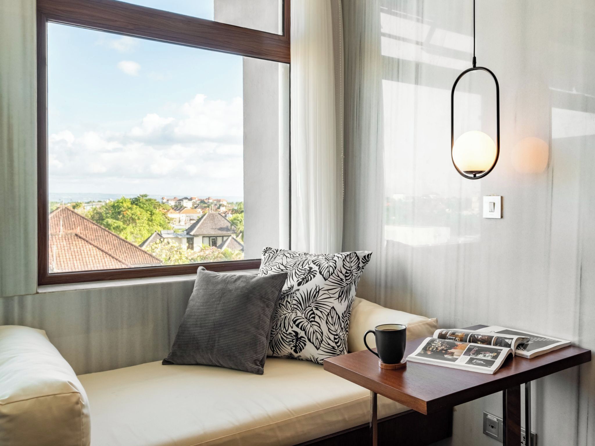Un rincón acogedor con vistas a un pueblo costero, una mesa con una revista y una taza, y una lámpara colgante.