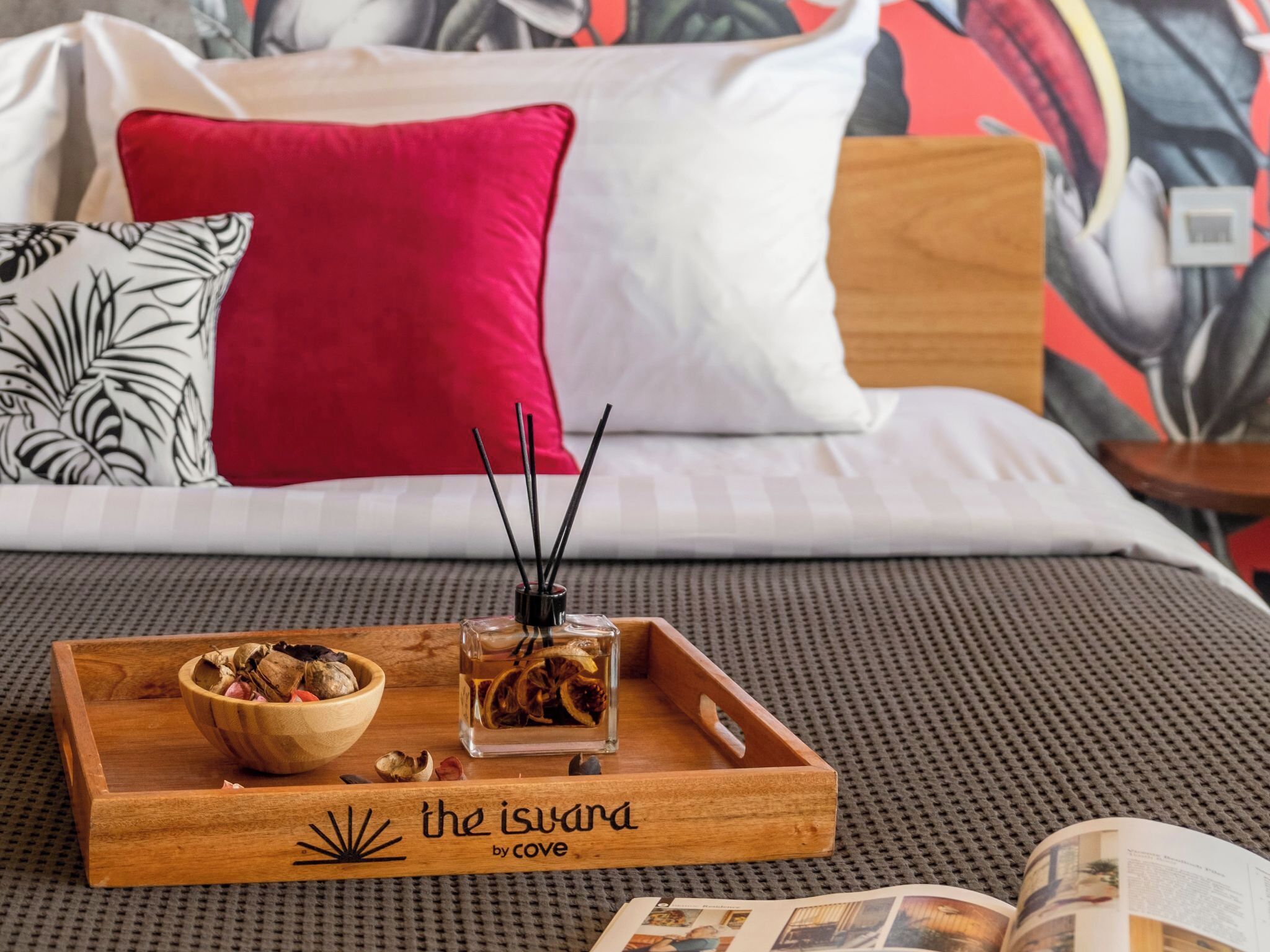 A neatly arranged wooden tray on a bed with a red pillow, a white pillow, and a black and white pillow. There are dried flowers in a glass container and a bowl of nuts on the tray. The bed has a grey textured bedspread and a wooden headboard.