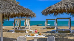 Un espacio para comer frente al mar, con sombrillas de paja, sillas de mimbre y una mesa con platos de comida y bebidas.