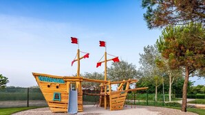 Una estructura de juego de barco pirata de madera con un tobogán, rodeada de grava y árboles.