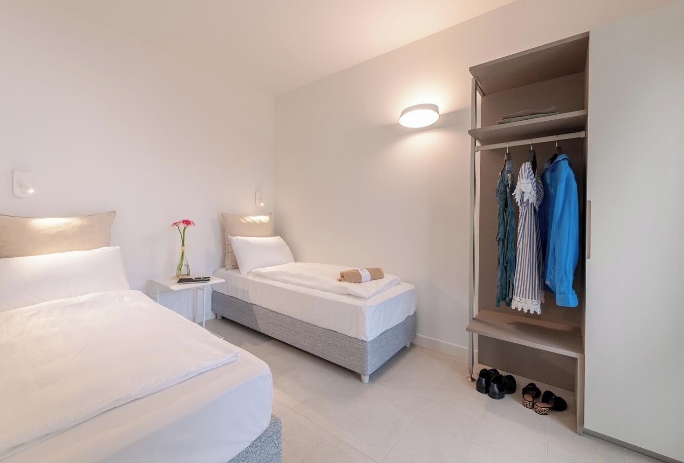 A modern hotel room with two beds, a bedside table with a vase of flowers, a wardrobe with hanging clothes, and a shoe rack.