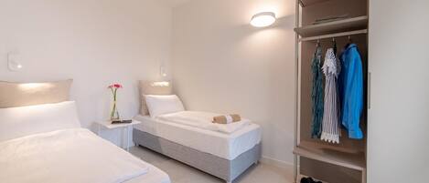 A modern hotel room with two beds, a bedside table with a vase of flowers, a wardrobe with hanging clothes, and a shoe rack.