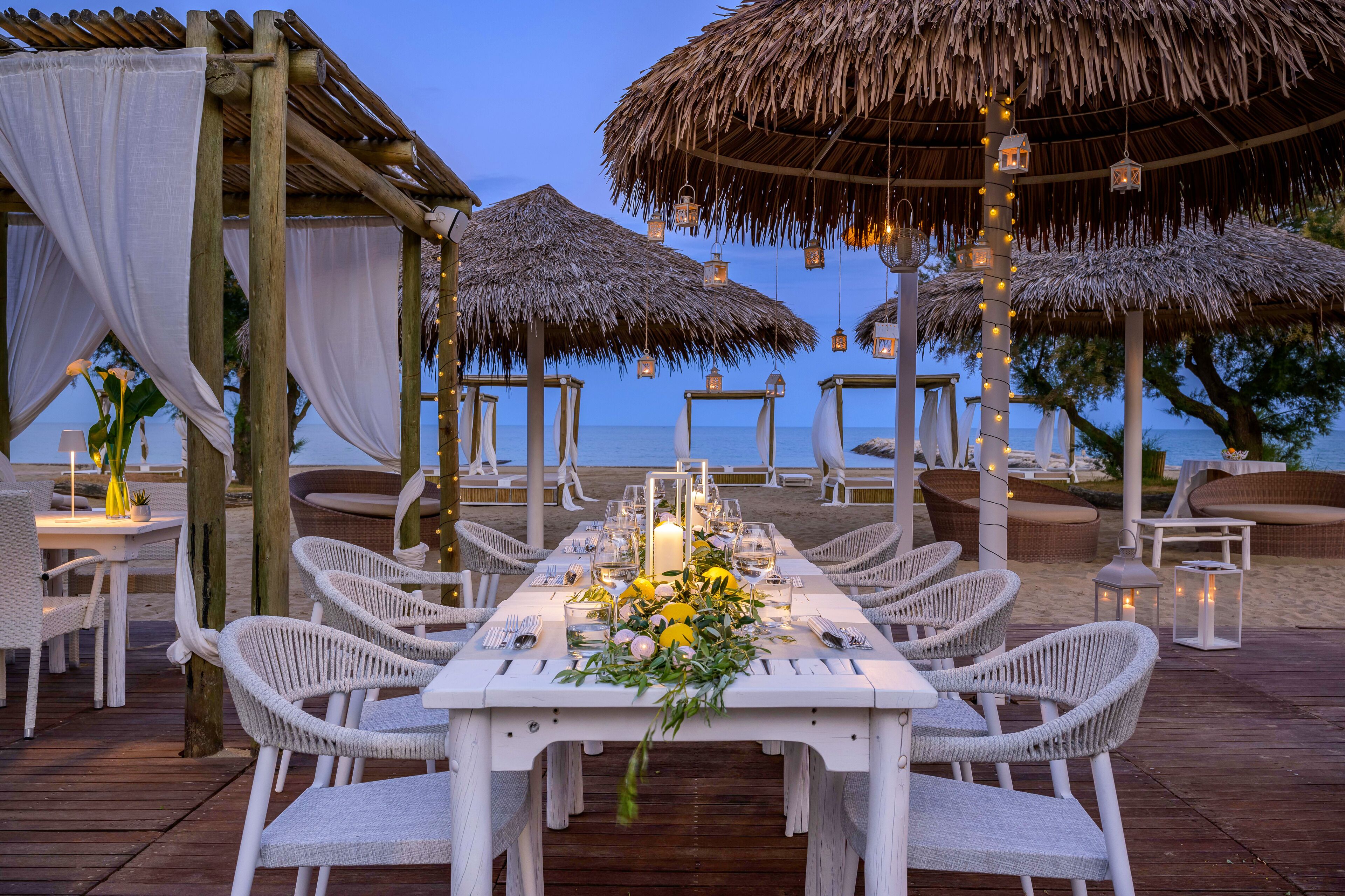 An outdoor dining area with white wicker chairs and tables, thatched umbrellas, and a view of the sea.