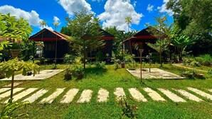 Un jardín tropical con dos cabañas de madera, rodeado de exuberante vegetación y un cielo azul despejado.