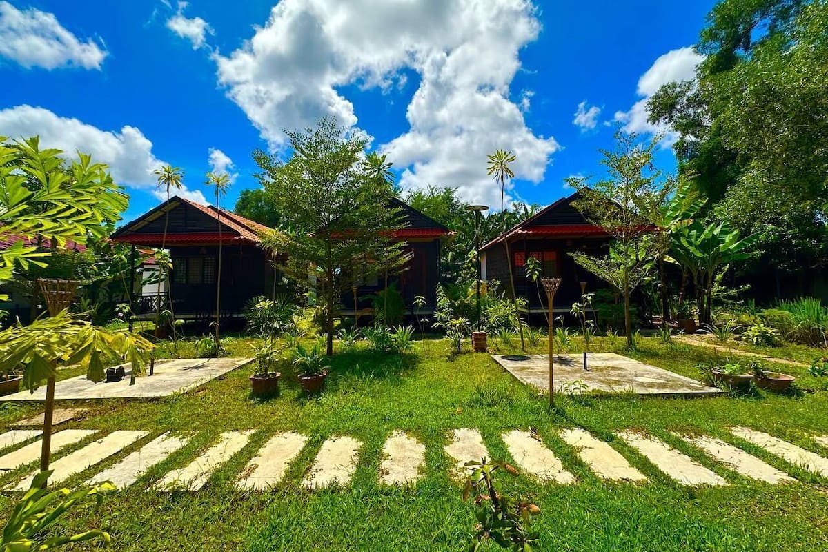 Un jardin tropical avec deux cabanes en bois, entouré d’une végétation luxuriante et surplombé d’un ciel bleu azur.
