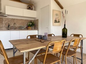 Un coin repas avec une table en bois et des chaises en osier, une cuisine avec des meubles blancs et un tableau accroché au mur.