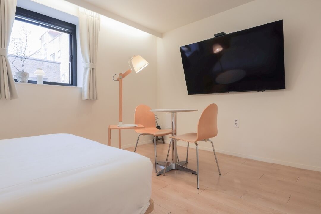 Habitación de hotel con cama, televisión montada en la pared, una mesita con dos sillas y una ventana con cortinas.