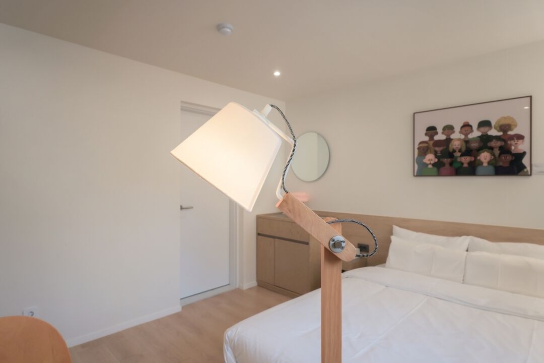 Una habitación de hotel con cama, mesita de noche, lámpara, un cuadro en la pared y una puerta.