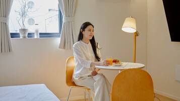 Ein Zimmer mit einem Bett, einem Esstisch mit sitzender Person, einer Lampe und einem Fenster mit Vorhängen.