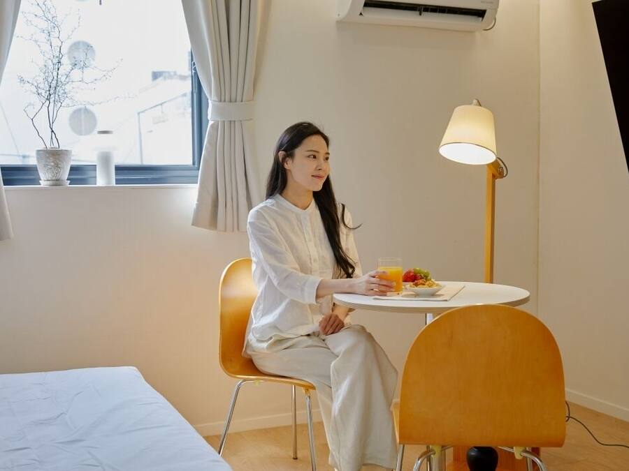 Ein Zimmer mit einem Bett, einem Esstisch mit sitzender Person, einer Lampe und einem Fenster mit Vorhängen.