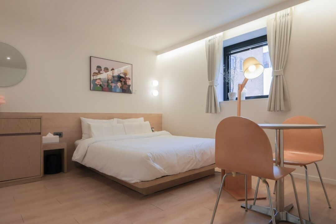 A hotel room with a bed, a bedside table, a chair, a lamp, a window with curtains, and a wall-mounted picture.