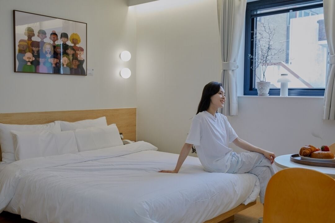 Una habitación de hotel con una cama, una mesa redonda con un plato de comida, una ventana con cortinas y una lámpara fijada a la pared.
