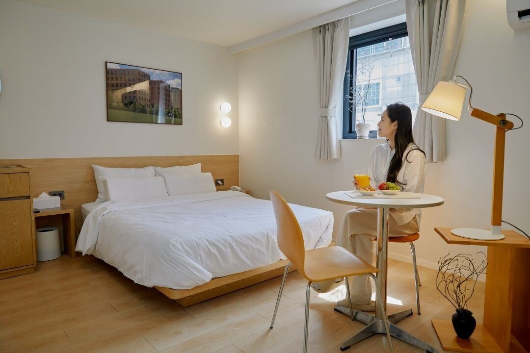 Una habitación de hotel con cama, escritorio, silla, lámpara y ventana con cortinas.