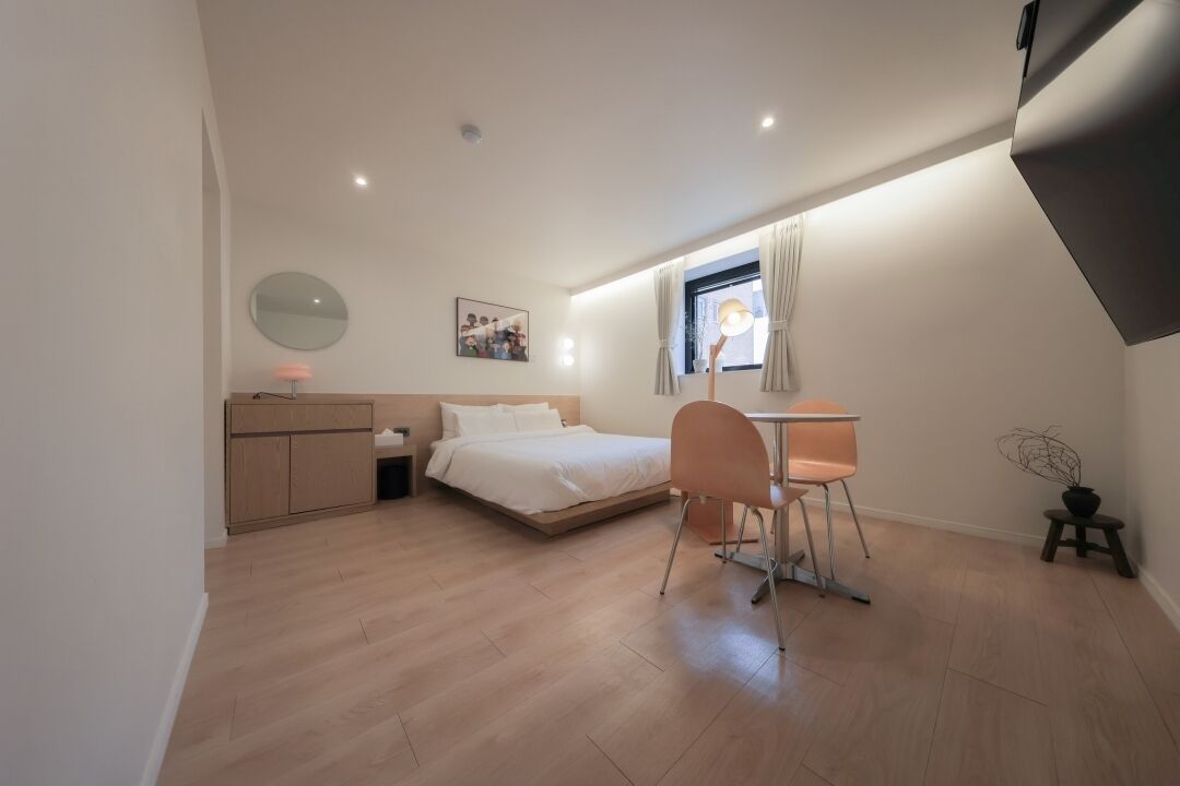 Un dormitorio con cama, un escritorio, una silla, un mueble pequeño, un espejo redondo, una ventana con cortinas y una planta en maceta.