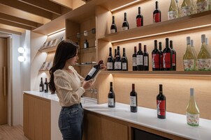 Una mujer observa botellas de vino en una barra de una tienda de vinos moderna y bien iluminada, con estantes de madera y una encimera blanca y limpia.