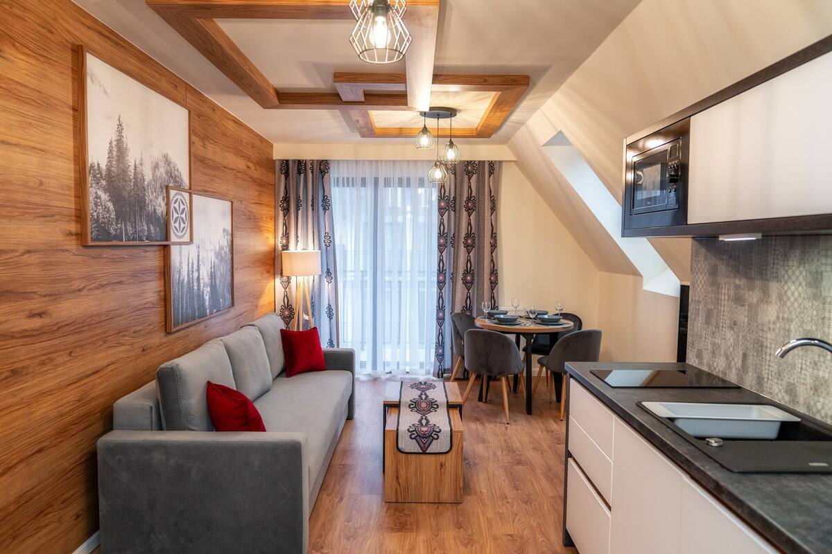 Sala de estar moderna com sofá, mesa de centro e área de jantar. Cozinha com micro-ondas e pia visível.