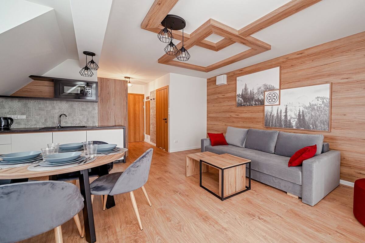 Sala de estar moderna com sofá cinza, mesa de centro de madeira e área de jantar com cozinha ao fundo.