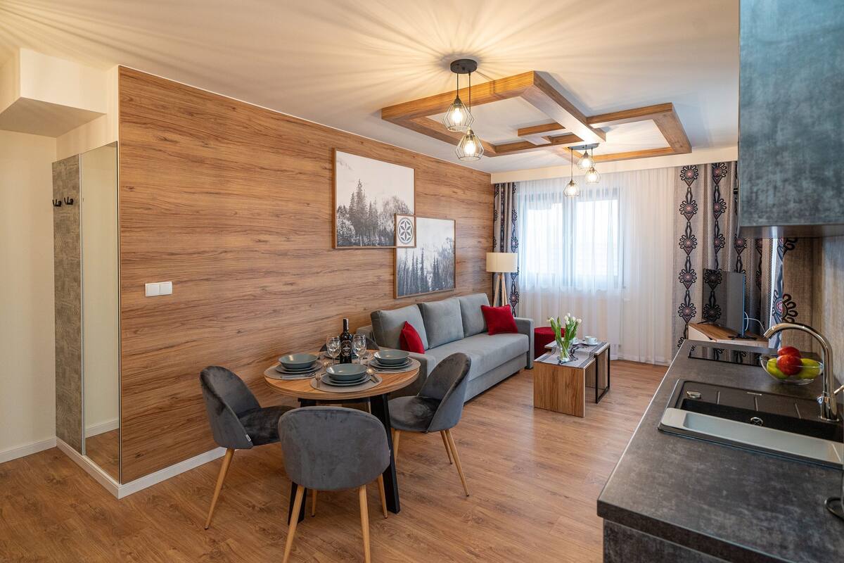 Uma sala de estar moderna com parede de madeira, um sofá, uma mesa de jantar e uma área de cozinha com pia e tigela de frutas.