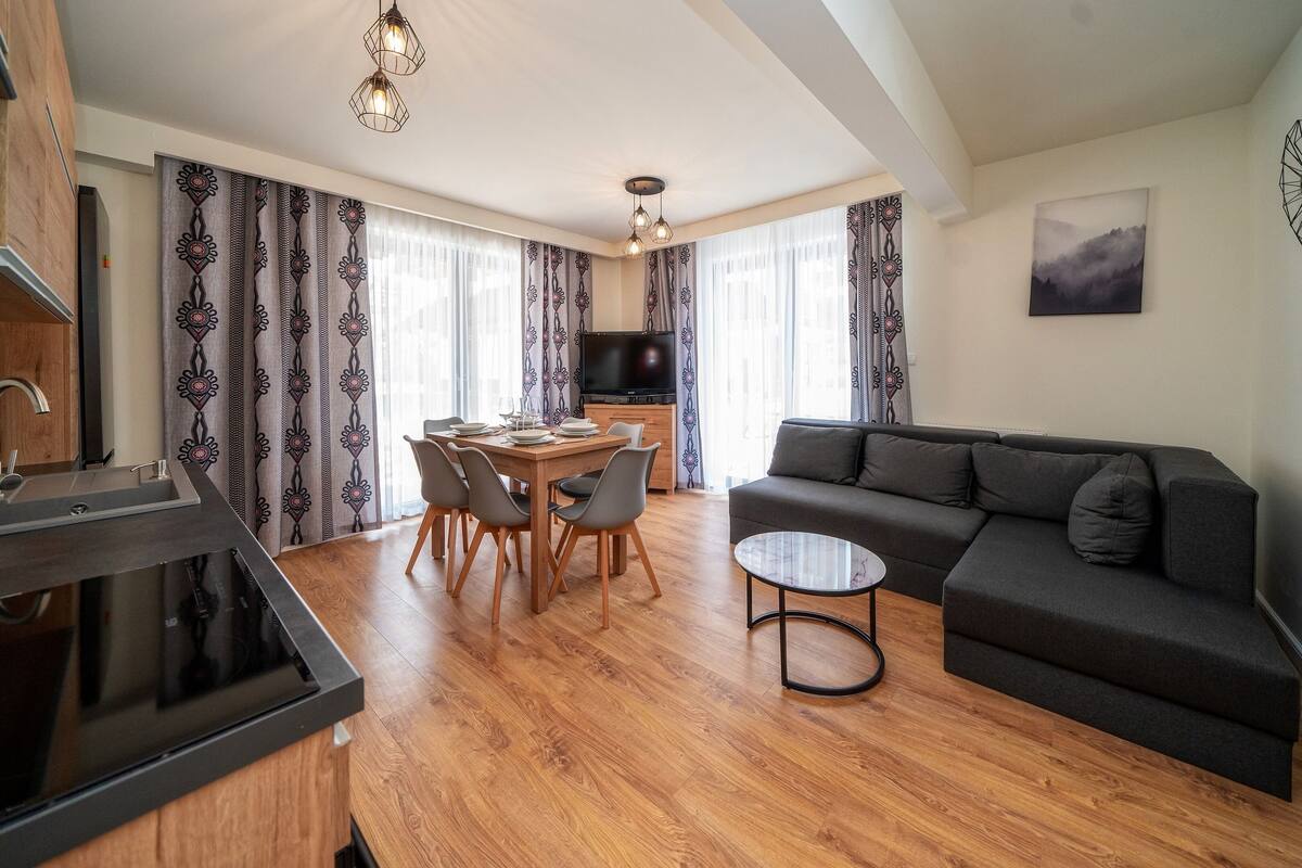 Uma sala de estar moderna com um sofá escuro, uma mesa de jantar com cadeiras, uma televisão e uma cozinha com fogão.