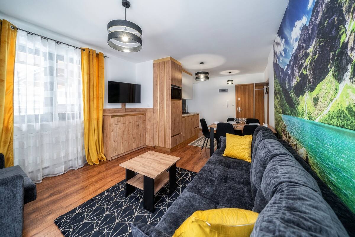 Uma sala de estar moderna com sofá, mesa de centro e televisão. Há uma área de cozinha com armários de madeira e uma mesa de jantar.