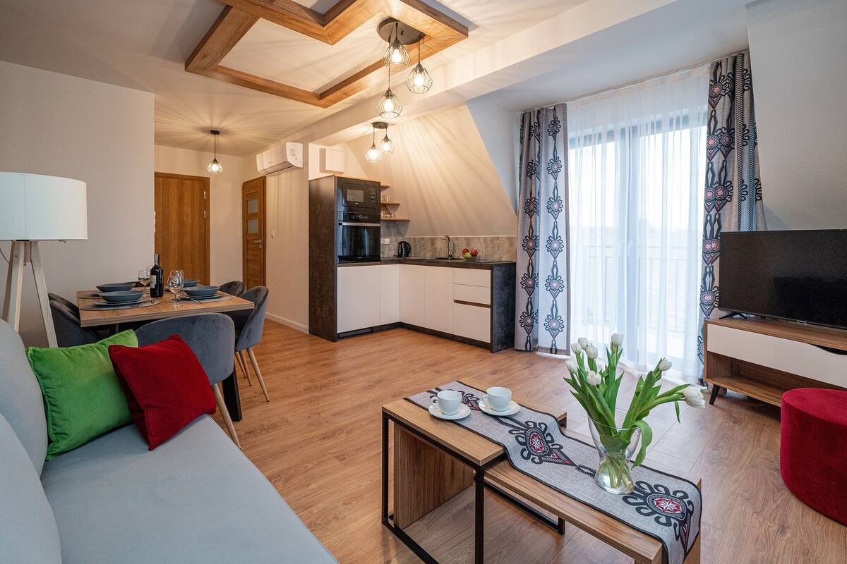 Uma sala moderna com sofá, mesa de jantar posta para dois, área de cozinha com TV e janela com cortinas.