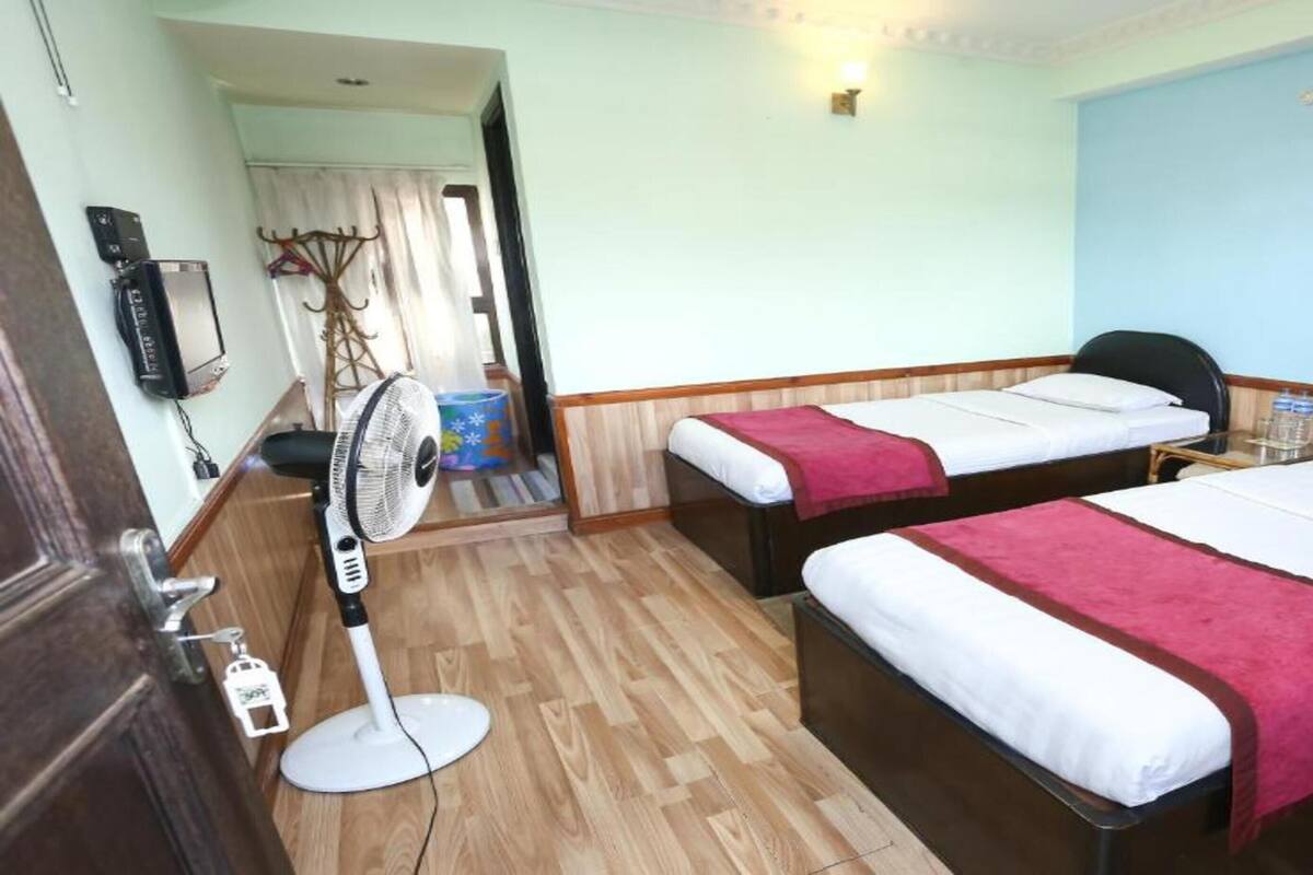 Habitación de hotel con dos camas individuales, piso de madera, ventilador de pie, mesita de noche con una botella de agua y televisor fijado a la pared.