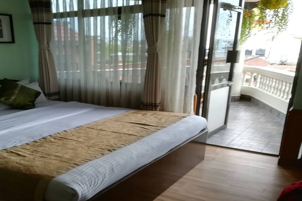 Un dormitorio con cama, almohadas, una colcha, una ventana con cortinas traslúcidas y un balcón con barandilla.