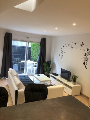 En moderne stue med sofa, sofabord, fjernsyn på et stativ og en balkon med udsigt over grønne områder.
