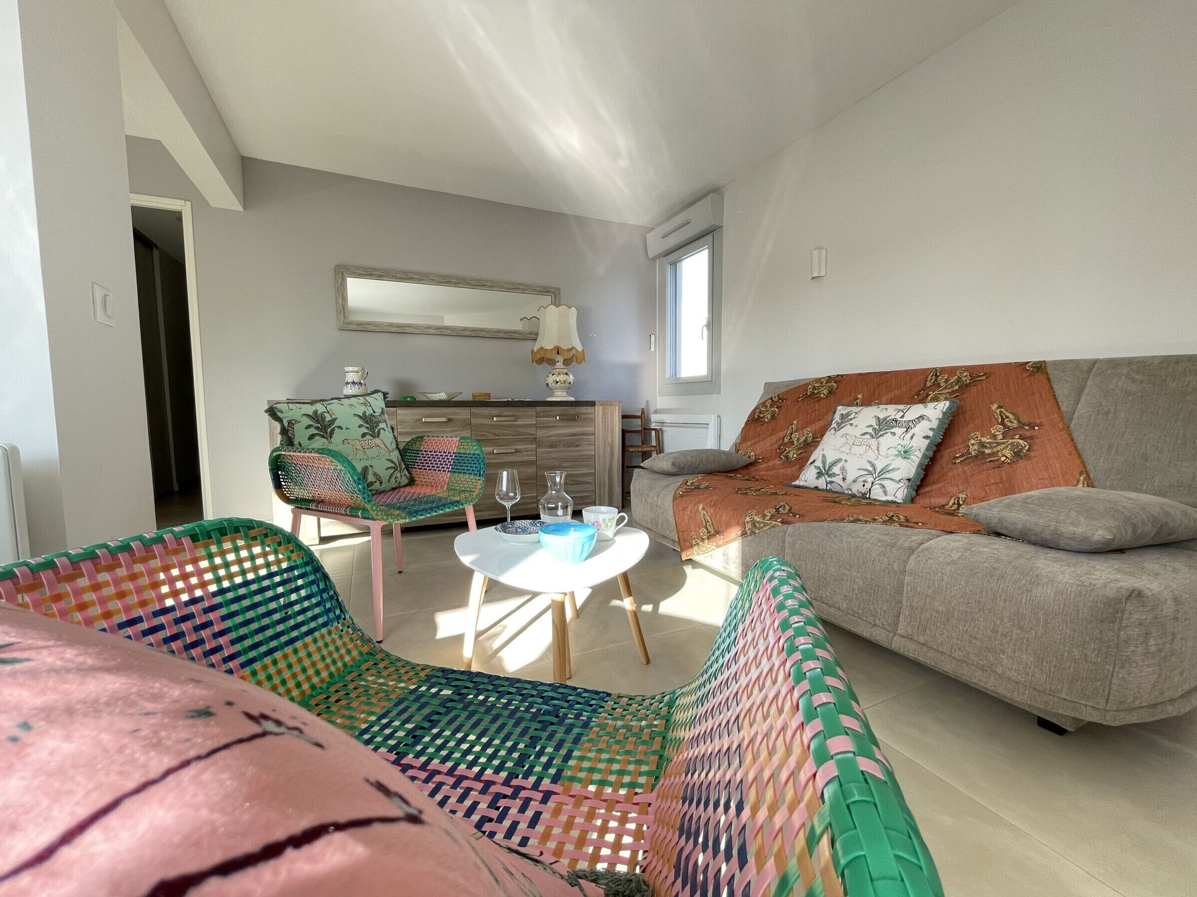 A modern living room with a sofa, a colorful woven chair, a small round table, and a kitchen area in the background.