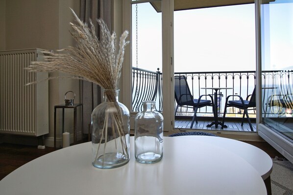 A room with a round table, a vase with pampas grass, and a glass bottle. There is a view of a balcony with chairs and a railing.