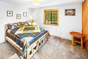 A bedroom with a wooden bed, a small wooden chair, a window with a view, and a wall-mounted picture. - Bear Mountain Log Lodge By Destination Big Bear: Walking distance to Bear Mountain,Golf Course and Hiking Trails! Hot Tub! Fenced Yard! Pet Friendly! (Big Bear Lake)