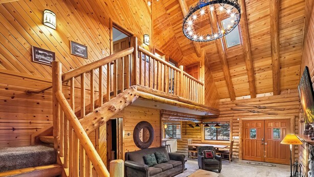 A log cabin interior with a wooden staircase, a cozy living area with a sofa and coffee table, and a dining area with a table and chairs.