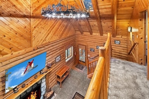 A log cabin interior with a fireplace, a television mounted on the wall, and a staircase leading to an upper level.