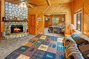 A log cabin interior with a stone fireplace, a large bed with a quilt, and a cozy sitting area.