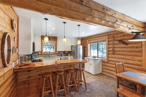 A rustic cabin interior with a wooden bar, stools, a dining table, and a kitchen area.