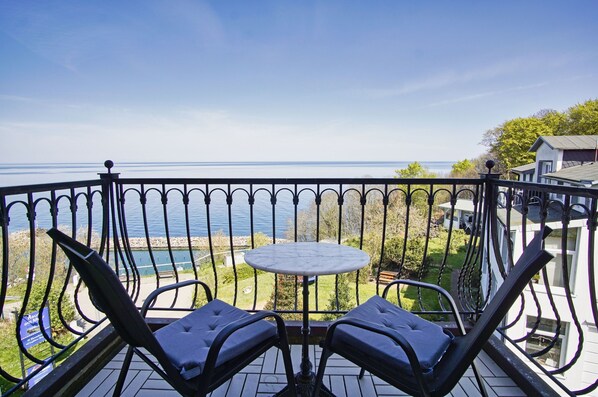 A balcony with a table and two chairs, overlooking a body of water and a grassy area with trees.