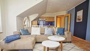 Una sala moderna con un sofá seccional color beige, una mesa redonda blanca y una cocina al fondo.
